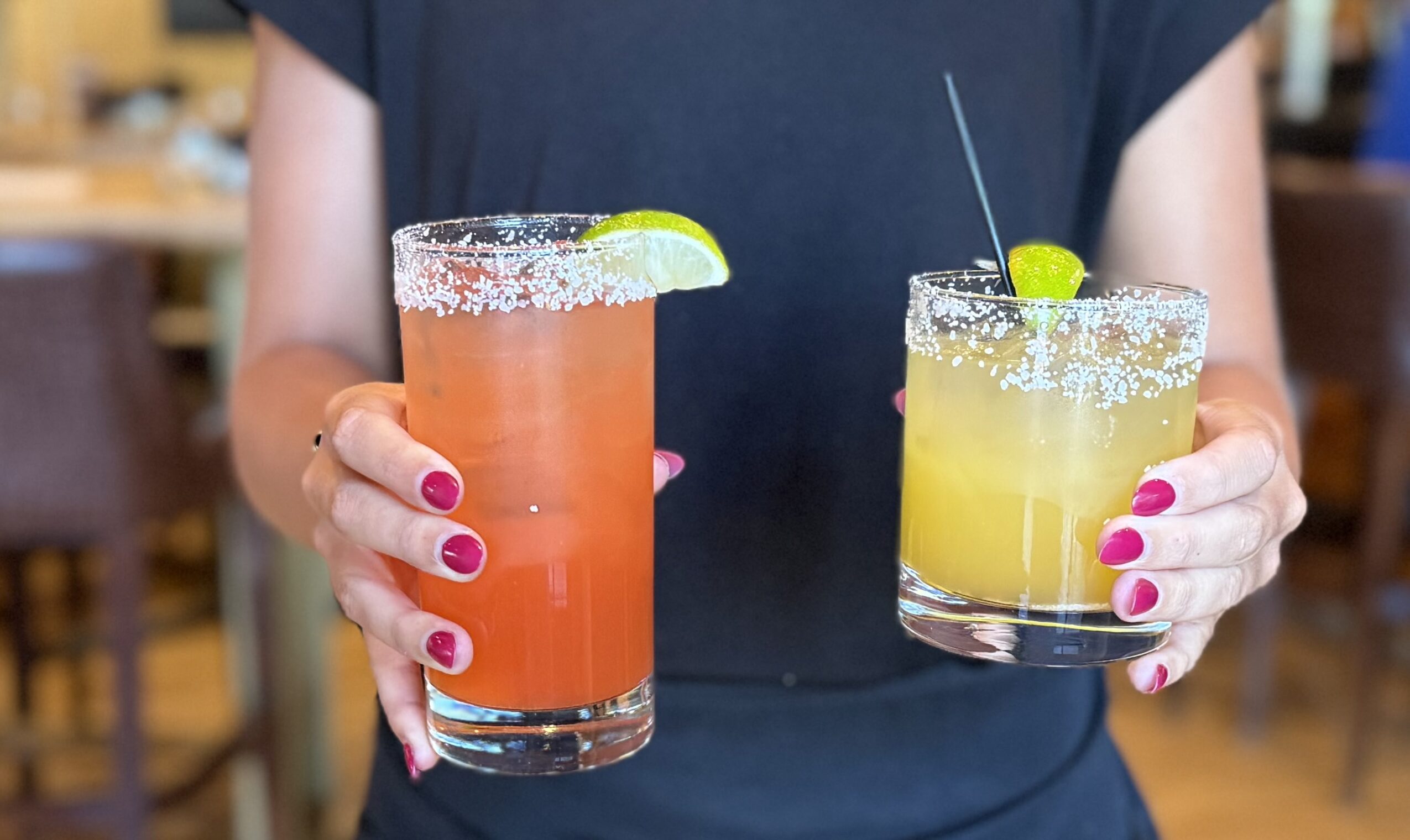 Person holding two colorful cocktails with lime garnishes and pink nail polish, in a restaurant setting.