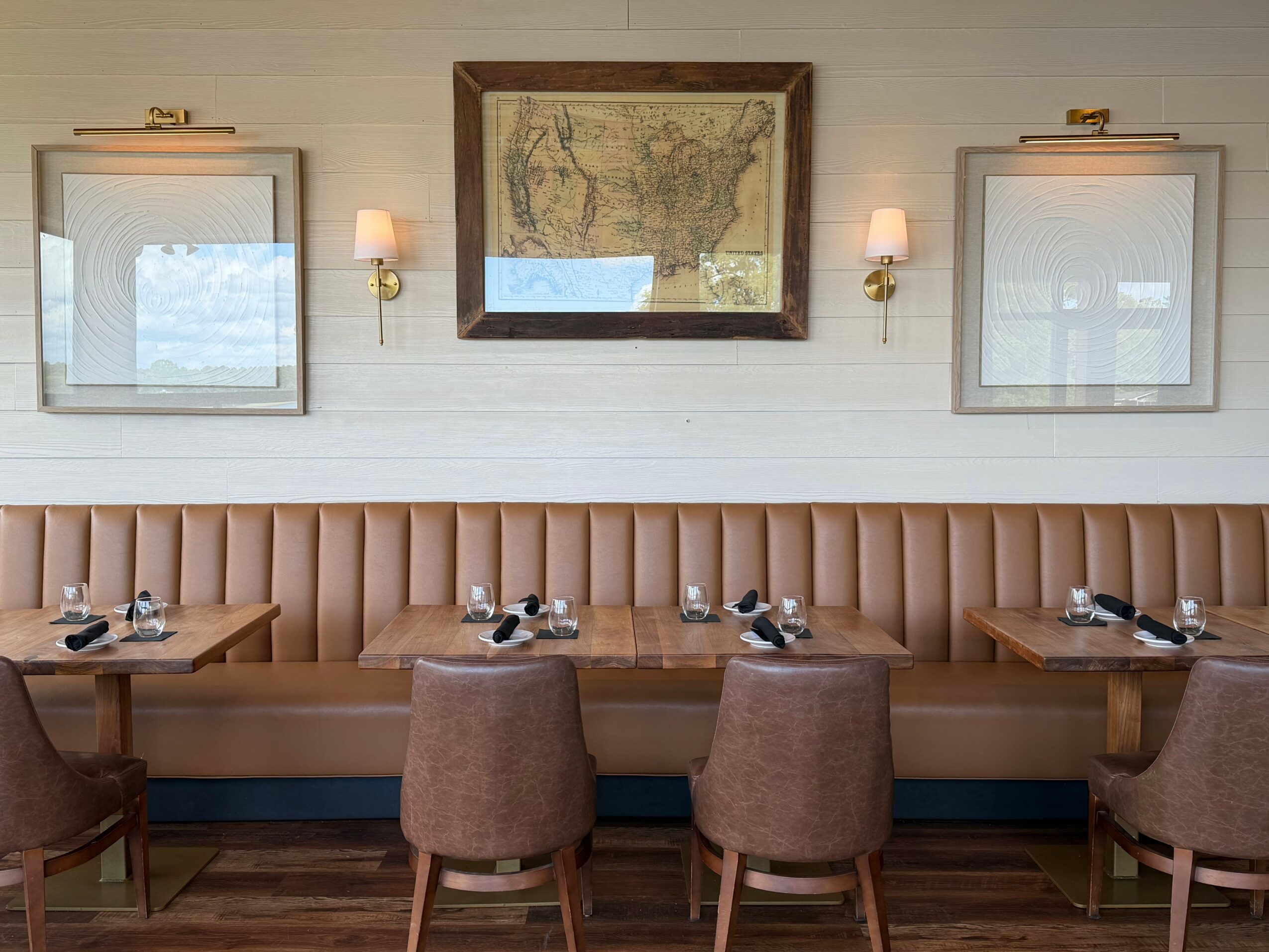 Dining area with a long beige upholstered bench, three tables with chairs, framed map on wall, and two wall lamps.