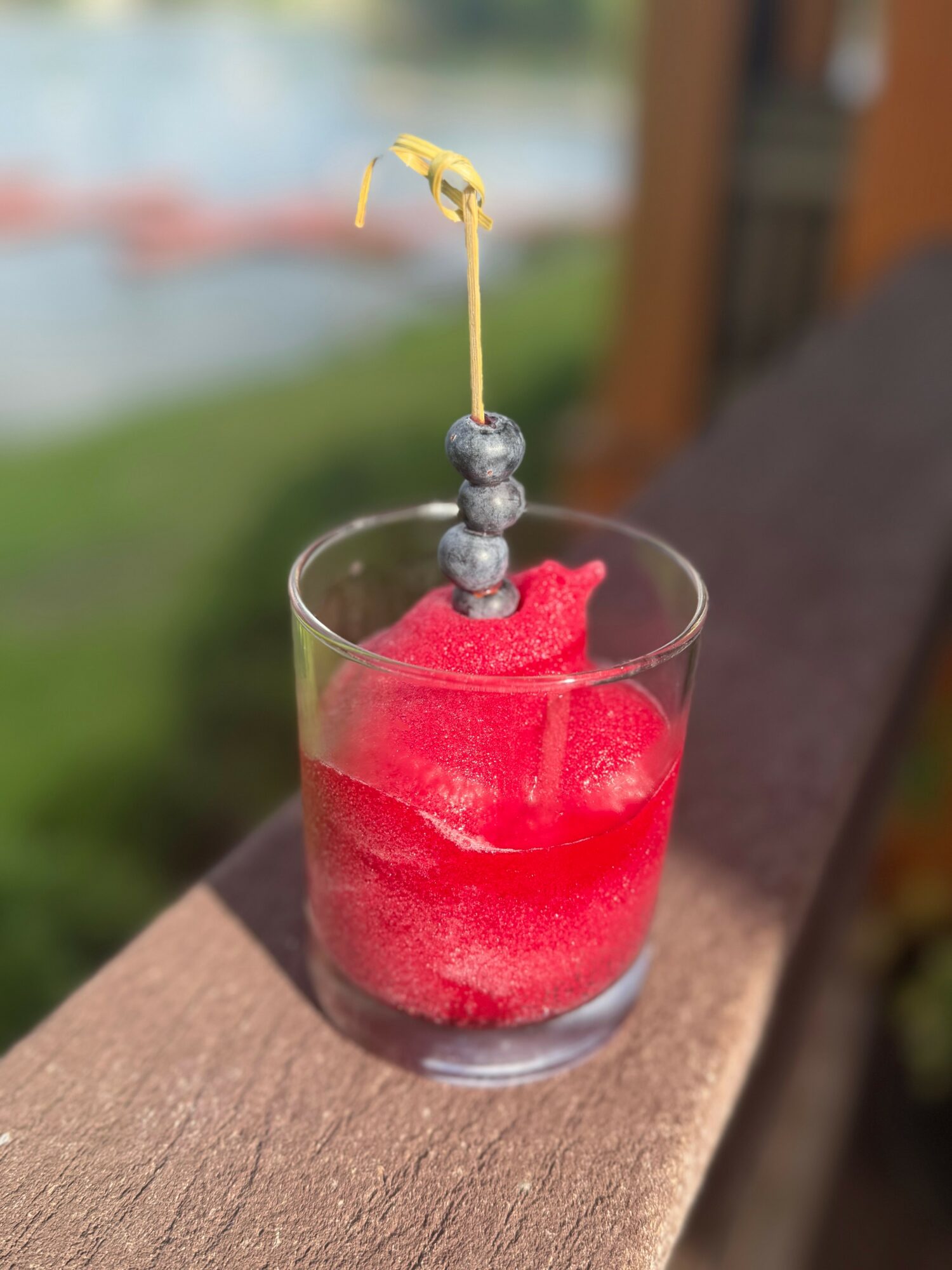 A glass with red frozen drink and a skewer with blueberries and a lemon peel, outdoors on a wooden surface.
