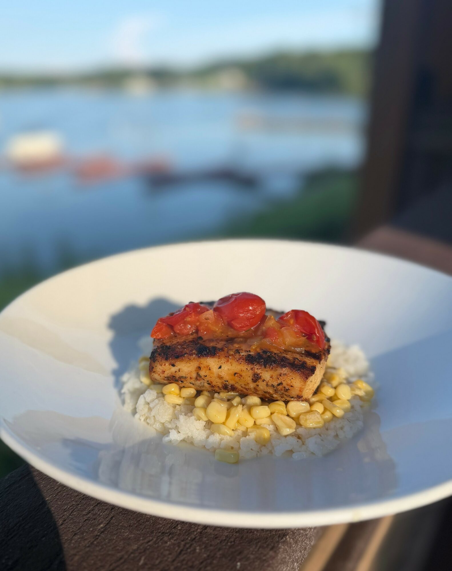 Slice of layered dessert with strawberries on top, served on a white plate with a blurred outdoor water scene in the background.