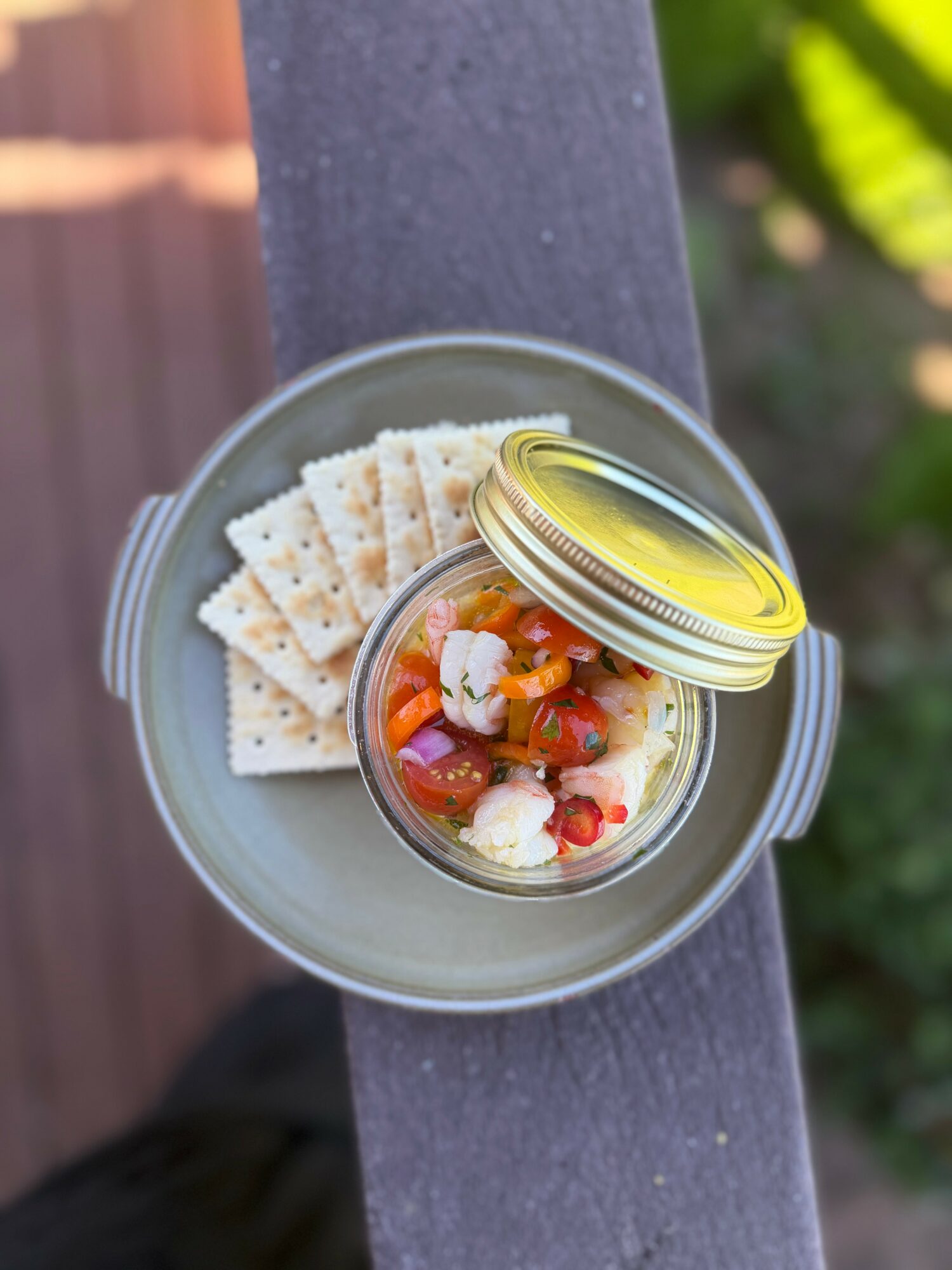 A jar of colorful salad with a gold lid, accompanied by crackers, on a gray surface outdoors.