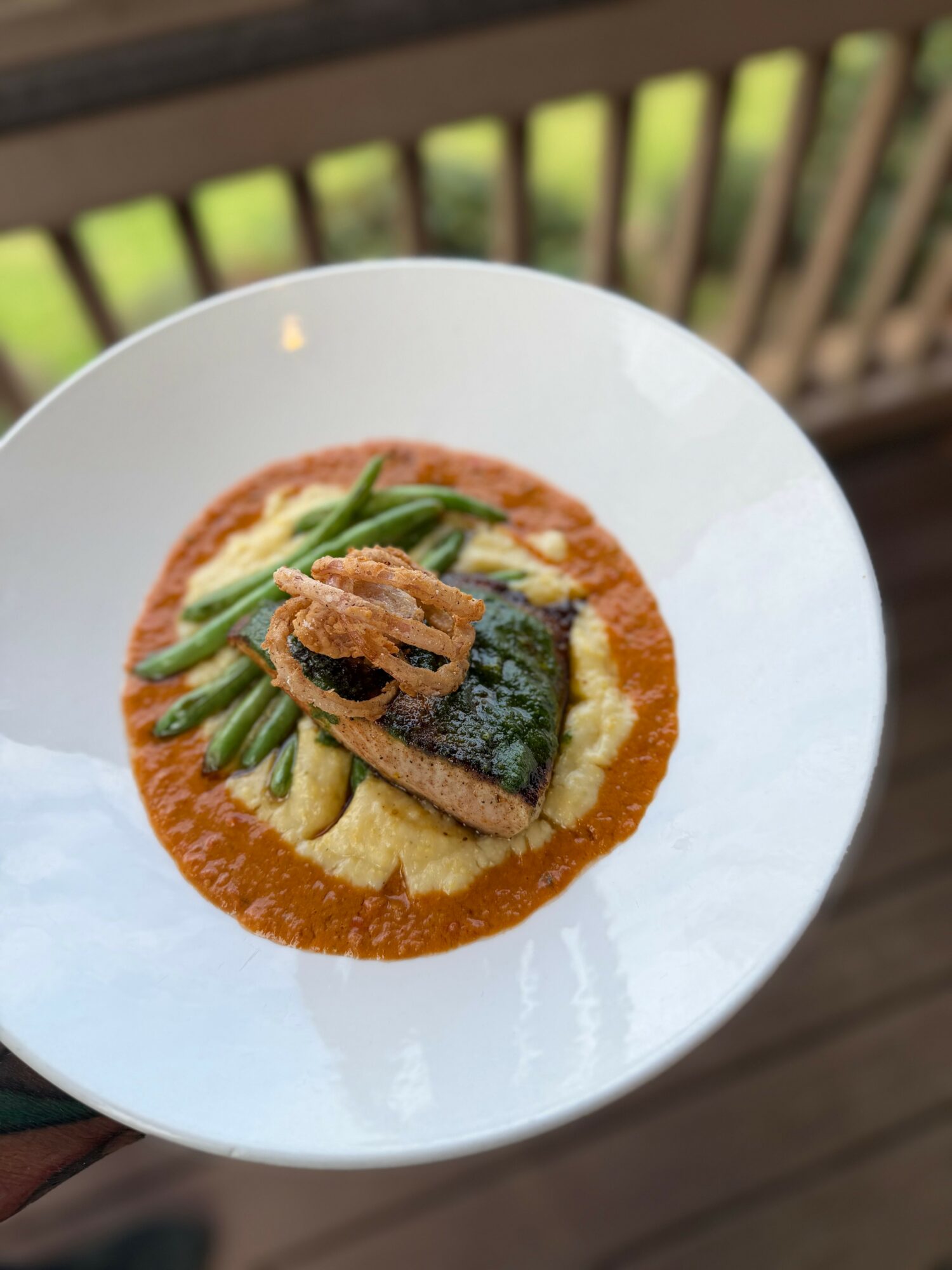 Plate with fish, green beans, and mashed potatoes in sauce, on a white dish outdoors, railing in background.