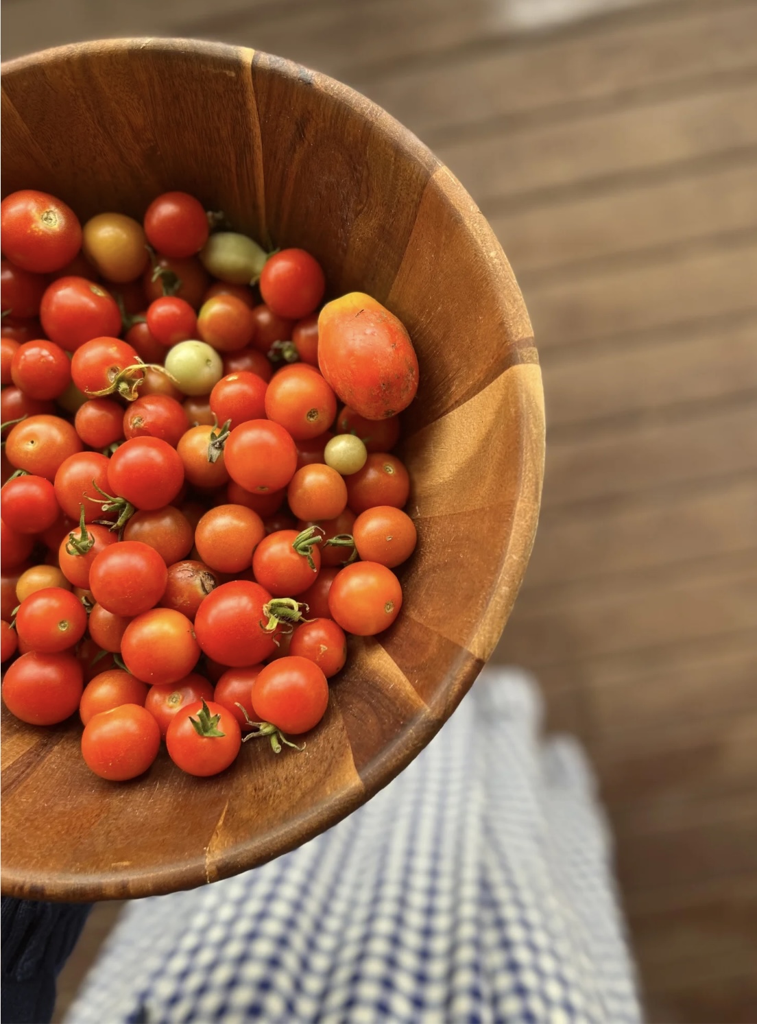 Wooden bowl filled with cherry tomatoes and a single apple, viewed from above, with a checkered cloth underneath.