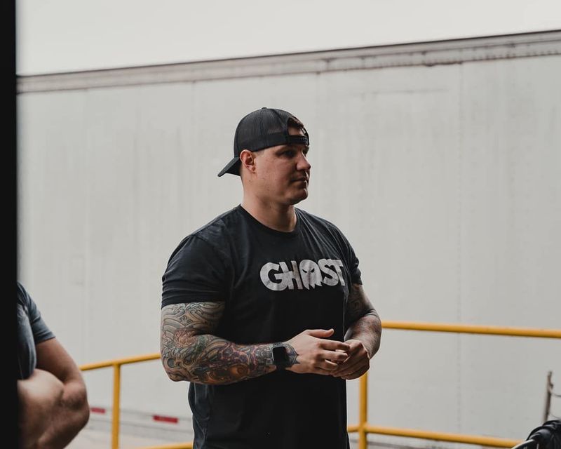 Man with tattoos wearing a black cap and T-shirt, standing outdoors near a yellow railing.