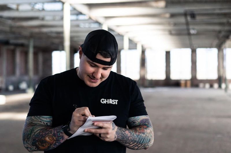 Young man with tattoos on arms, wearing a black cap and shirt, writing on a notepad in an empty industrial space.