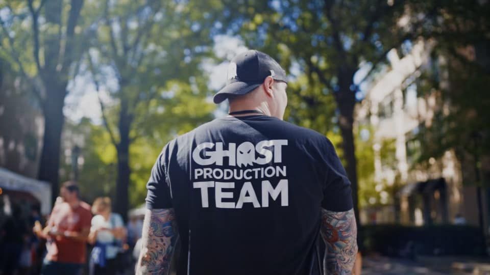Person wearing a black shirt with white text, outdoors with trees and buildings in background.