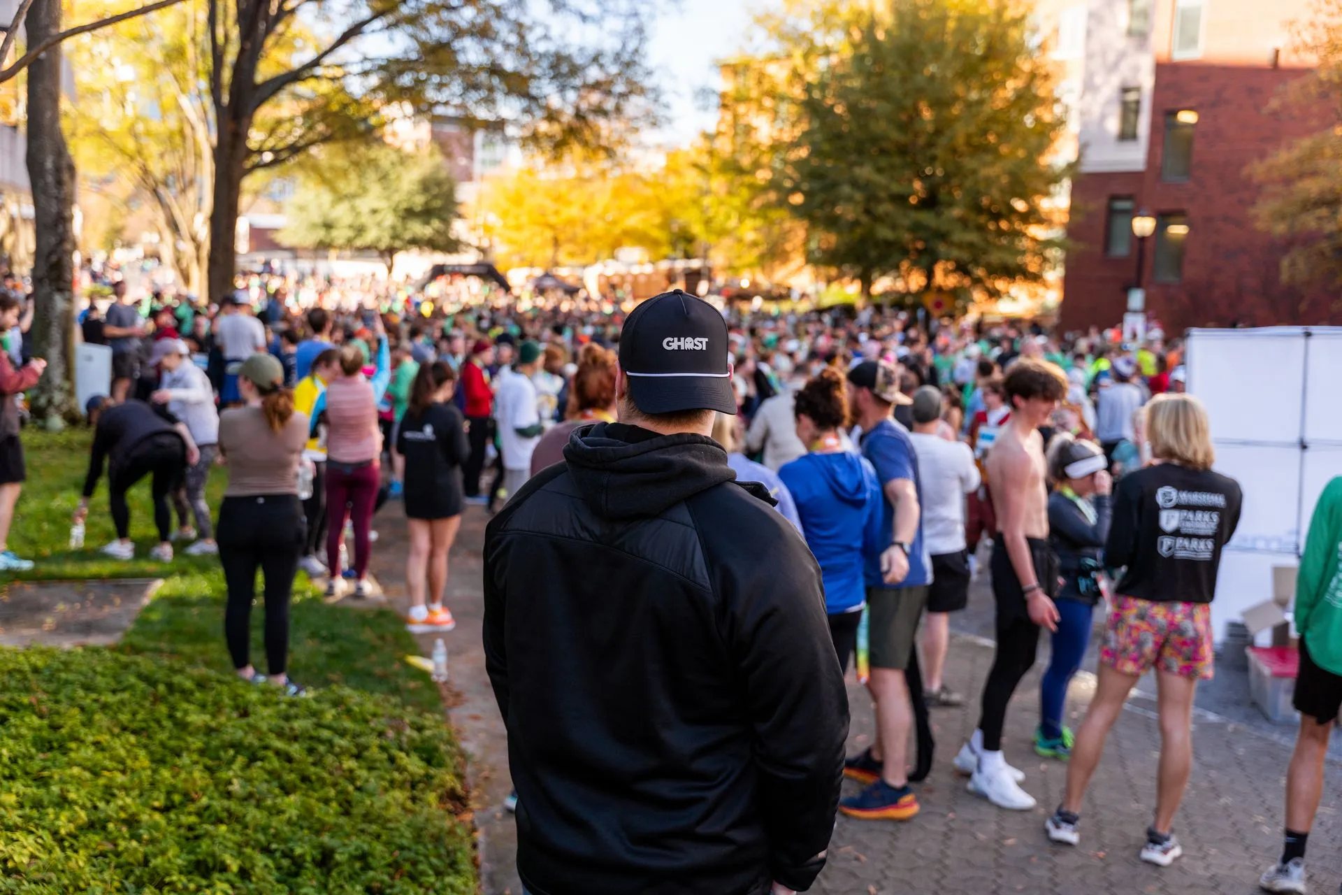 Crowd of people gathered outdoors in a park with trees and buildings in the background.
