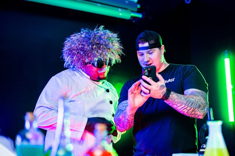 Two people looking at a phone, one with curly hair and a chef's coat, the other with tattoos and a cap, in a neon-lit setting.