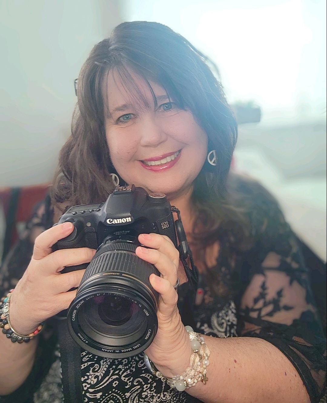 Woman smiling holding a camera, wearing earrings and a bracelet, with a bright background.