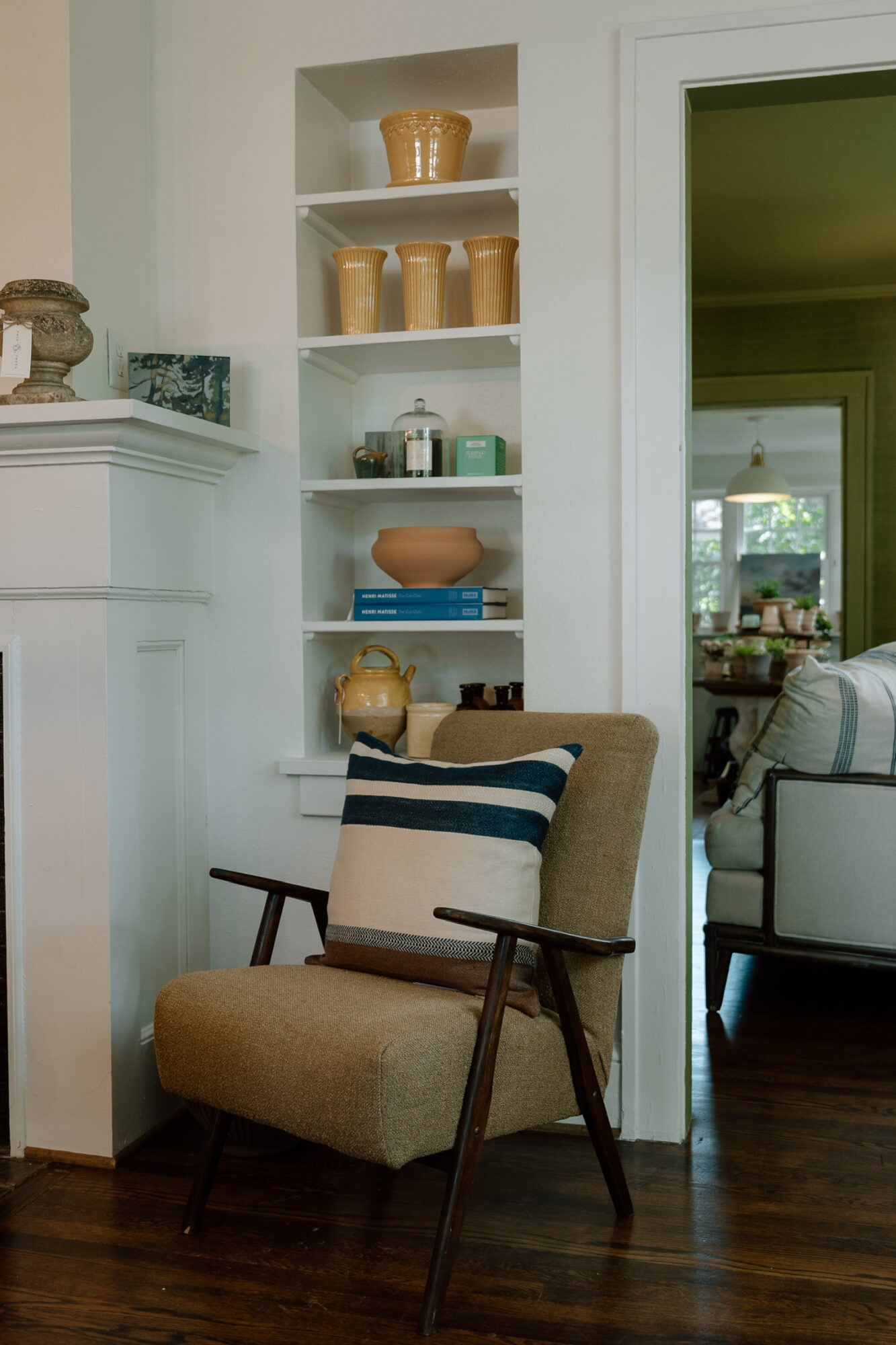 Living room corner with a beige armchair, white built-in shelves, and a doorway to another room with a sofa.