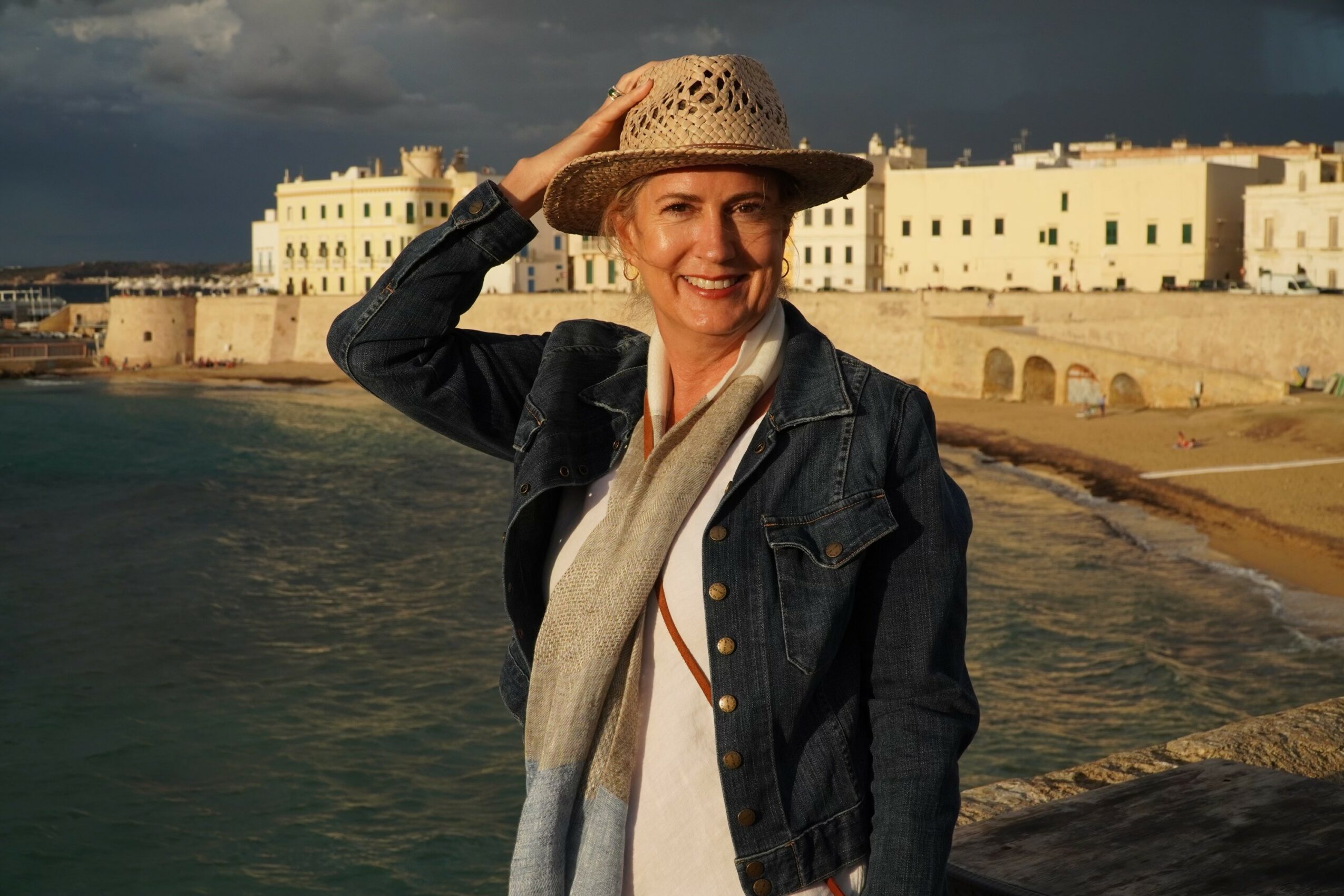 Woman smiling, wearing a hat and denim jacket, standing near water with buildings in the background.