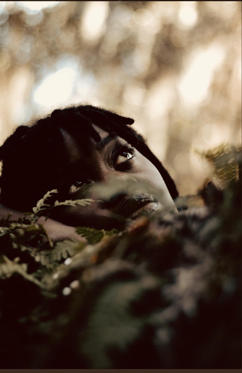 Person with dreadlocks partially hidden among foliage, looking upward with a blurred outdoor background.