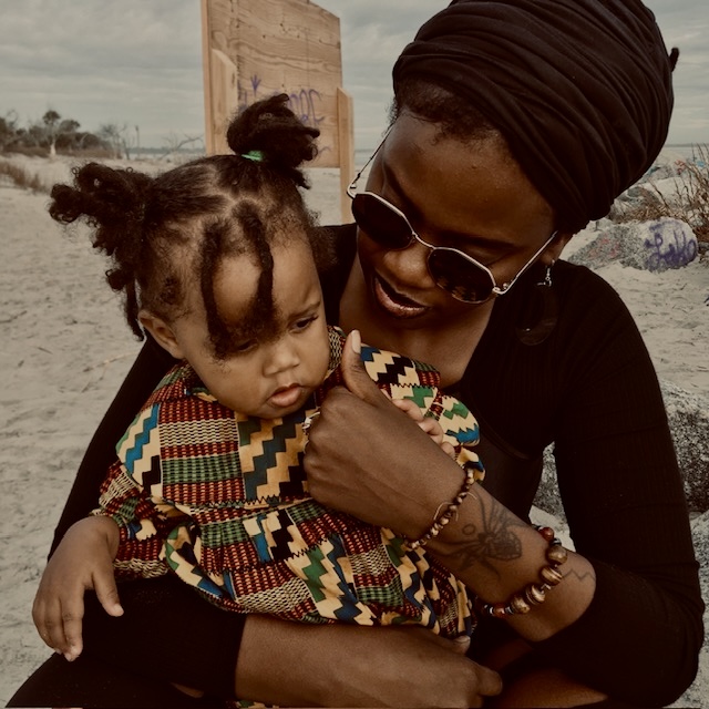 Woman holding a child outdoors, both wearing colorful clothing, woman with sunglasses and headwrap, child with pigtails, sandy background.