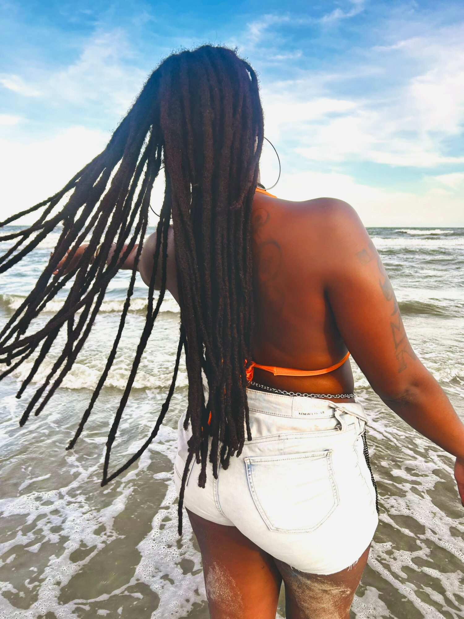 Person with long dreadlocks standing on the beach facing the ocean, wearing white shorts, with a blue sky and waves.