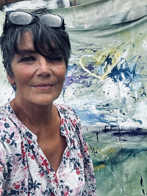 Woman with short dark hair and glasses on her head, smiling, wearing a floral top, standing in front of an abstract painting.