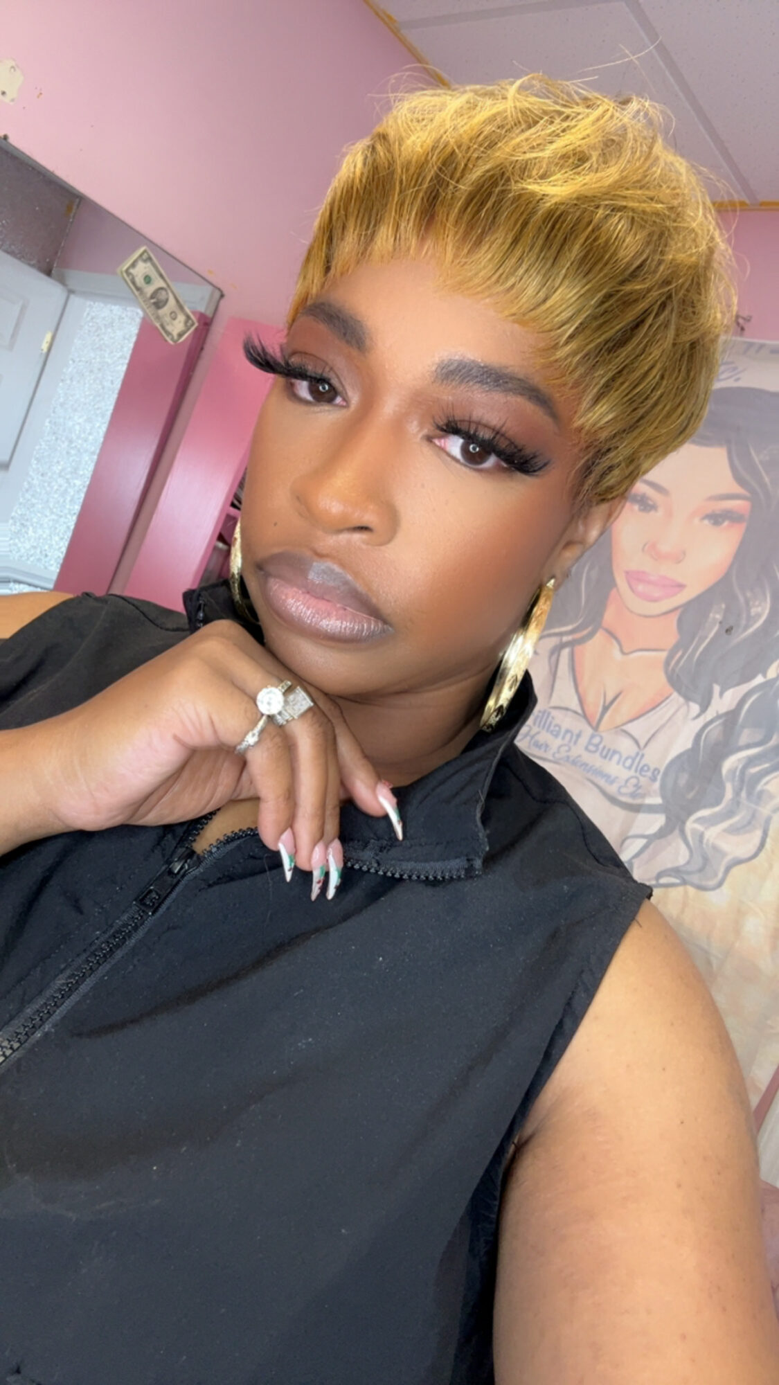 Woman with short blonde hair, makeup, and earrings, wearing a black sleeveless top, in a room with pink walls.