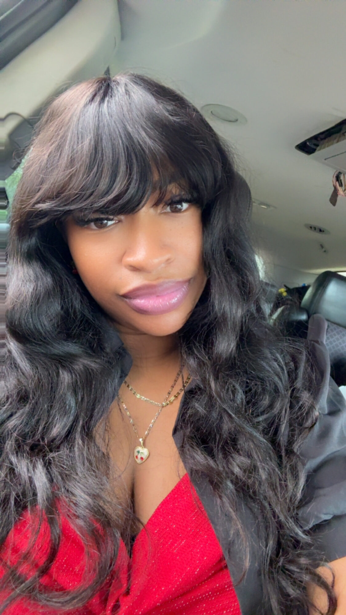 Woman with long wavy hair and bangs, wearing a red top and gold necklace, inside a vehicle.