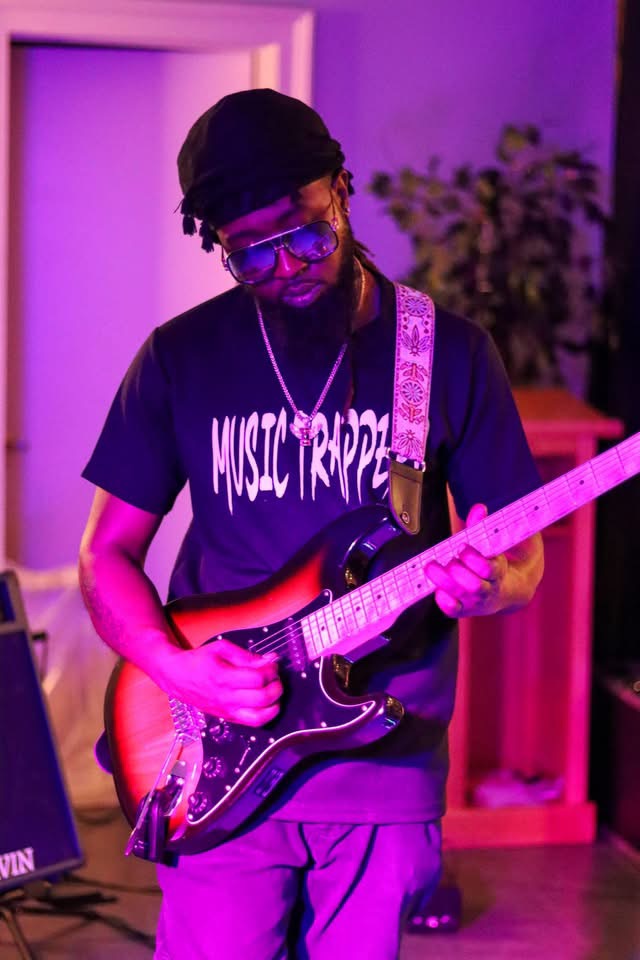 Person playing electric guitar in a room with purple lighting, wearing glasses, a black cap, and a black T-shirt.