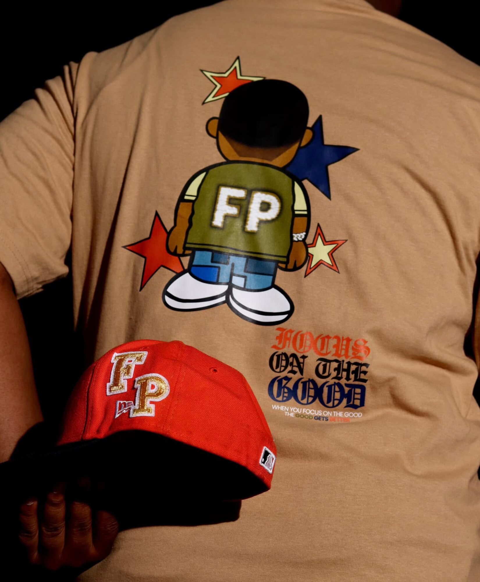 Person wearing a beige shirt with a cartoon character and text, holding a red cap with white lettering.