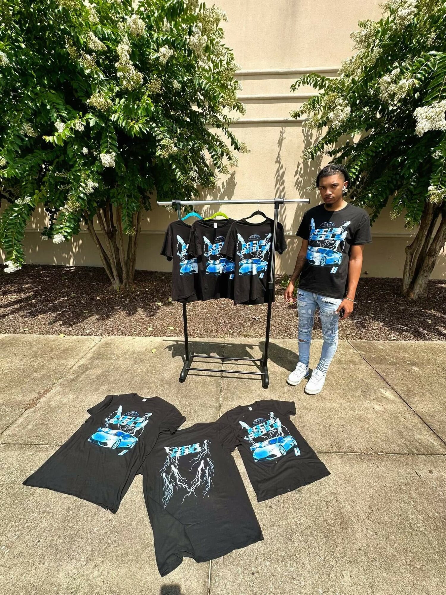 Young person standing outdoors next to a display of T-shirts with a blue and black design, surrounded by flowering bushes.
