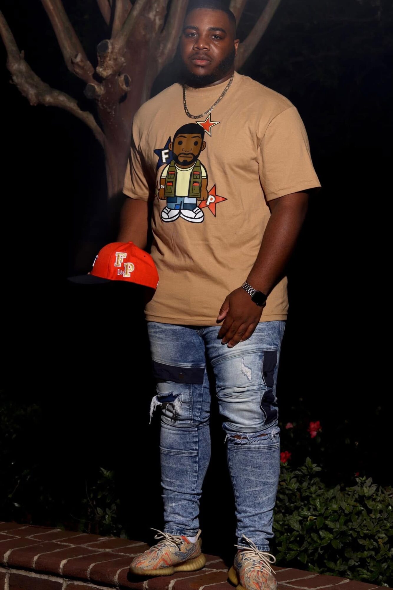 Man standing outdoors at night, wearing a beige T-shirt with a cartoon character, ripped jeans, and sneakers, holding a red cap.