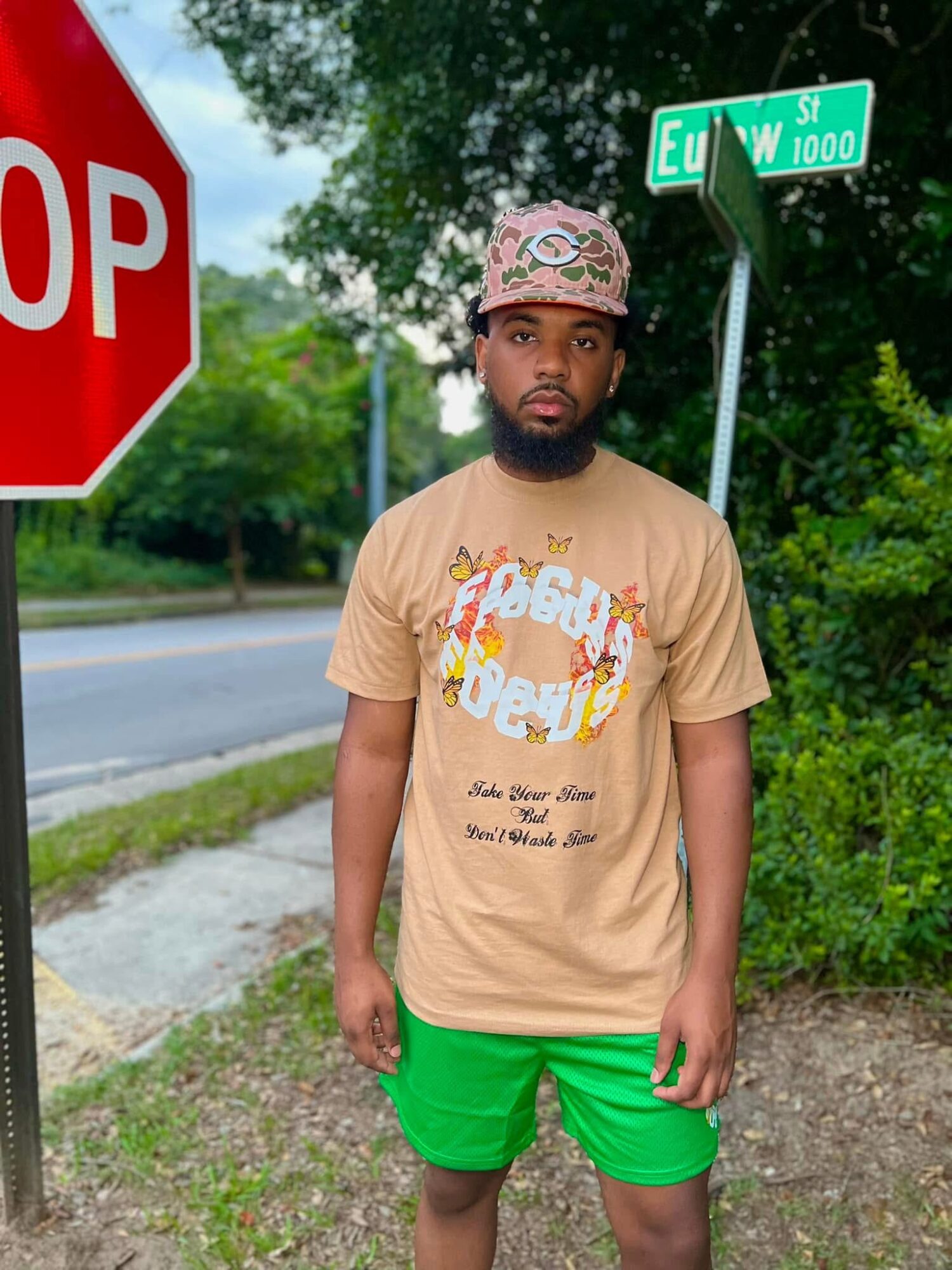 Young man with a beard standing outdoors near a stop sign and street signs, wearing a beige T-shirt, green shorts, and a patterned cap.