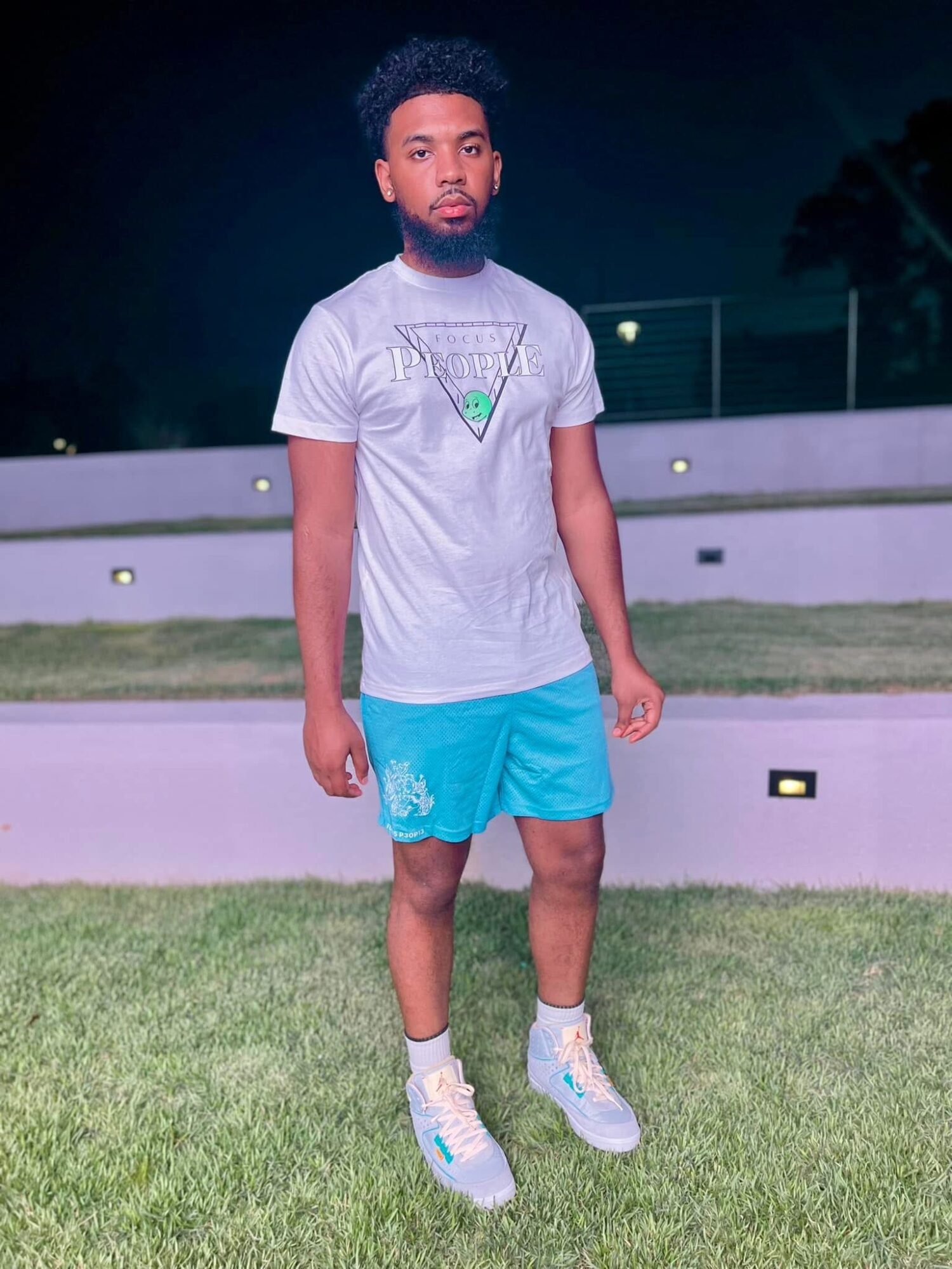 Young man standing outdoors at night on grass, wearing a gray t-shirt, blue shorts, and sneakers.