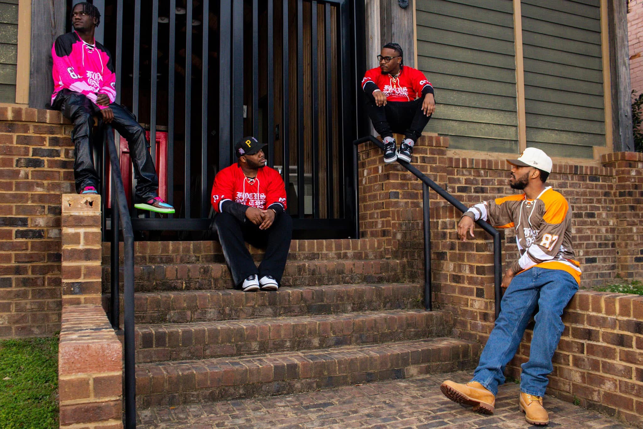 Four people sitting and standing on steps outside a building, two wearing red shirts, one in pink, one in yellow and white, holding a microphone.