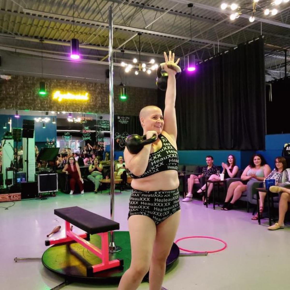 A performer with a bald head and a black and gold costume stands on stage, holding a kettlebell and raising one arm, with an audience watching.