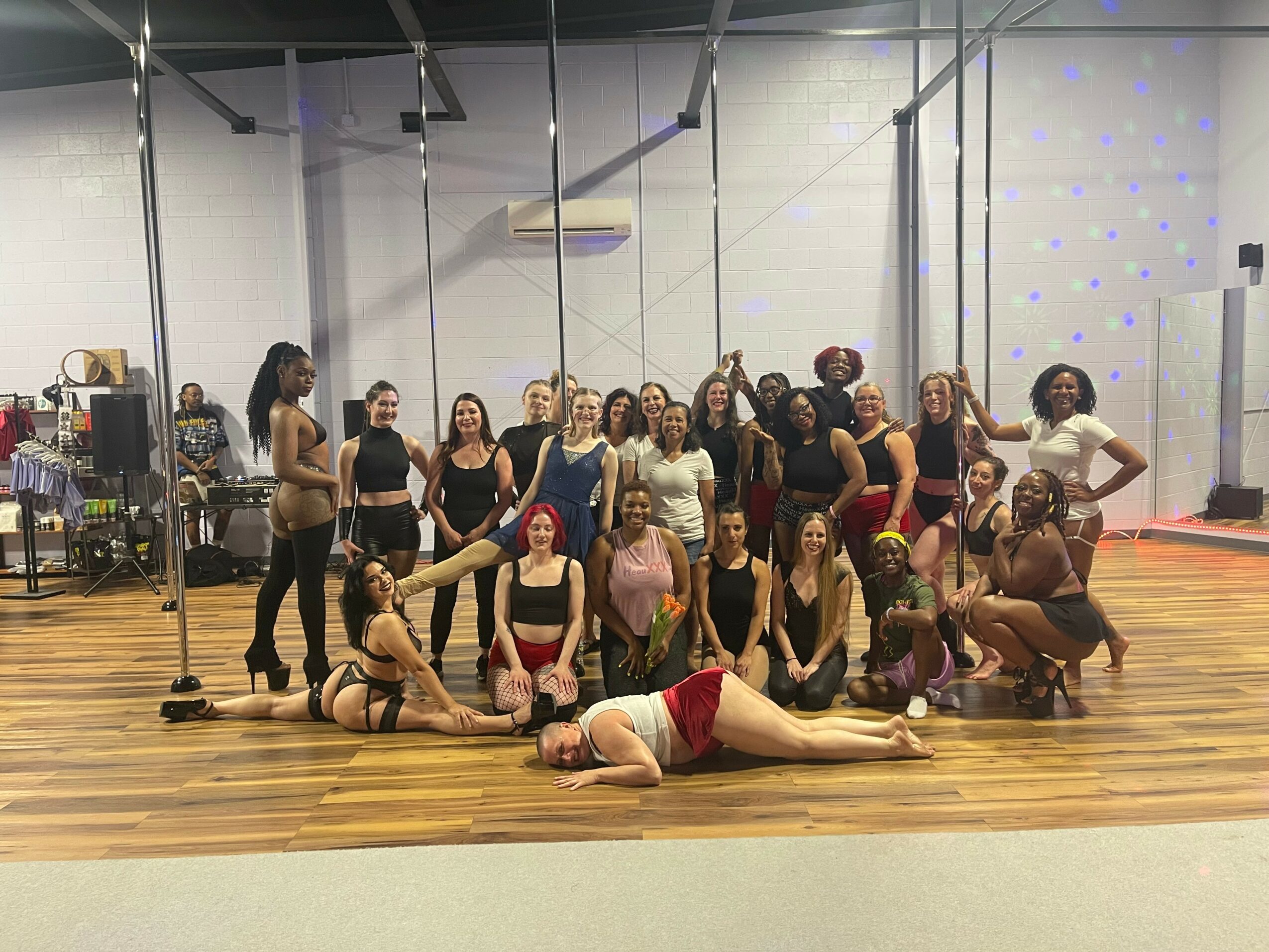Group of women in a dance studio, some sitting, some standing, with a person lying on the floor in front.