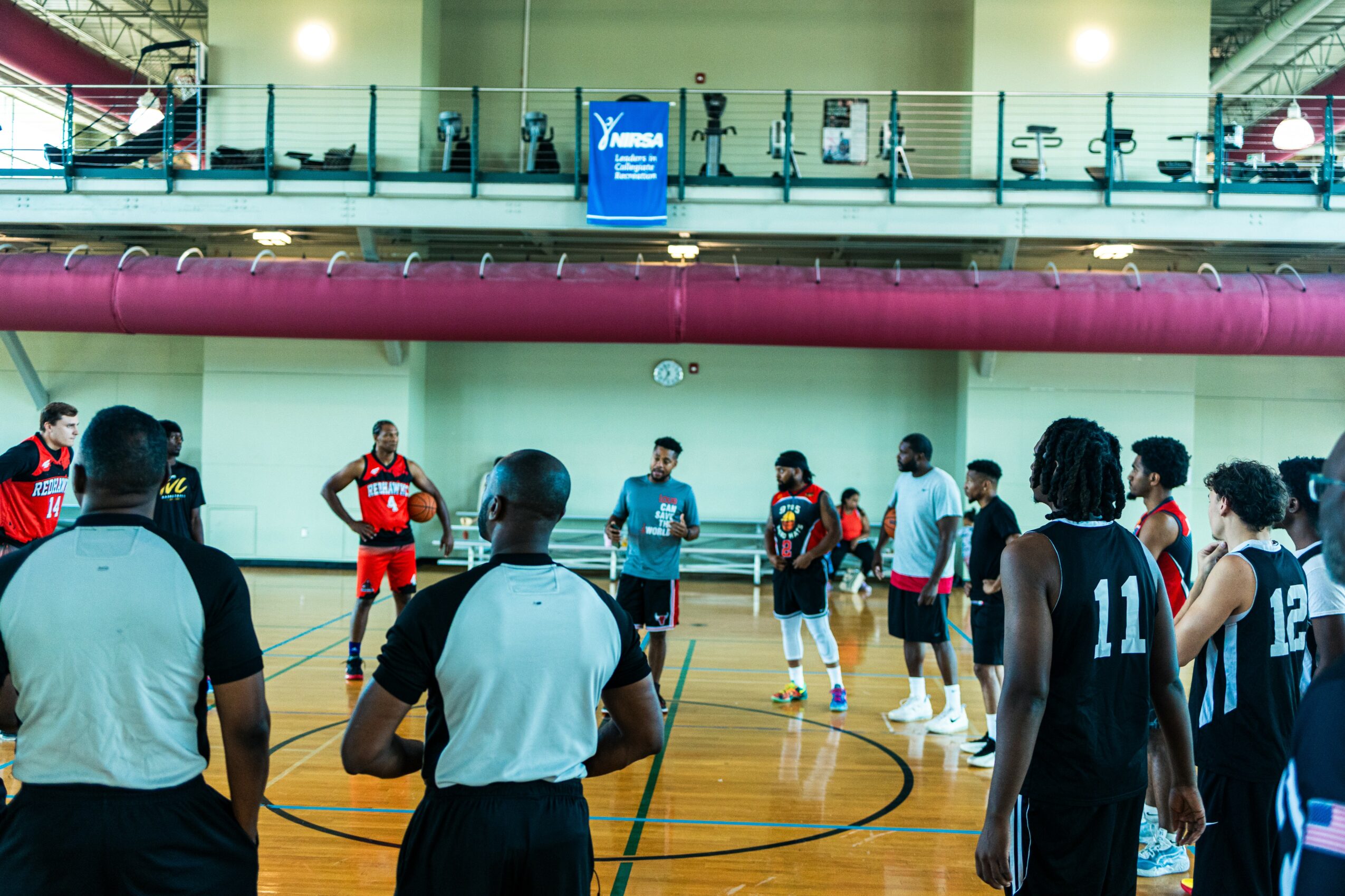 Indoor basketball game with players and referees on court, spectators on upper level, and a clock on wall.