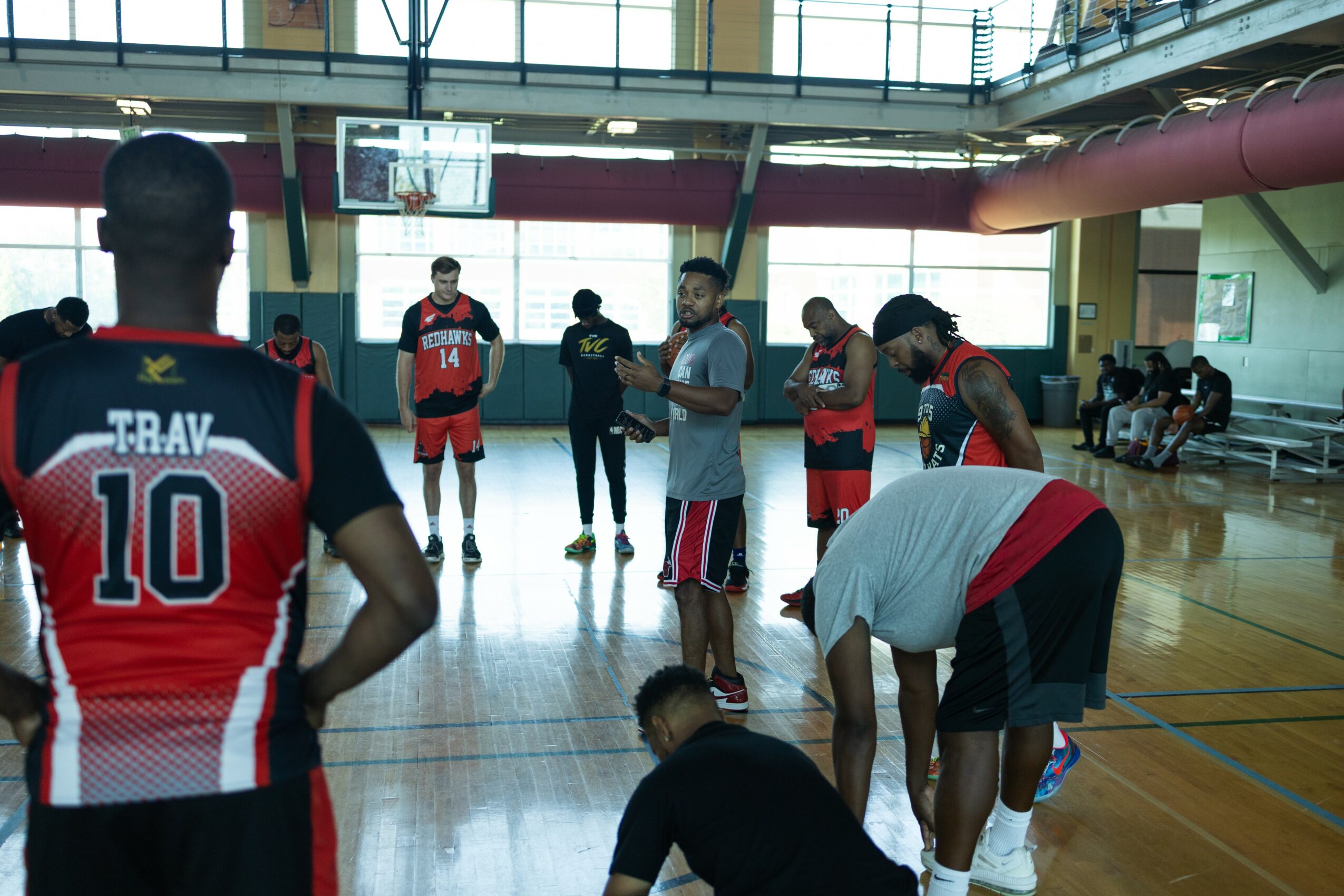 Group of people in a gymnasium, some wearing sports uniforms, standing and sitting near a basketball court.