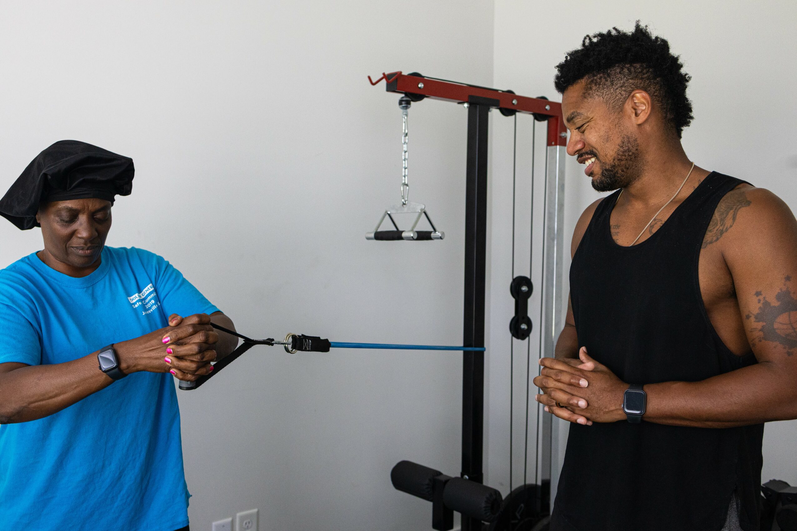 Two people in a gym, one woman adjusting a resistance band, and a man smiling with hands clasped, standing near exercise equipment.