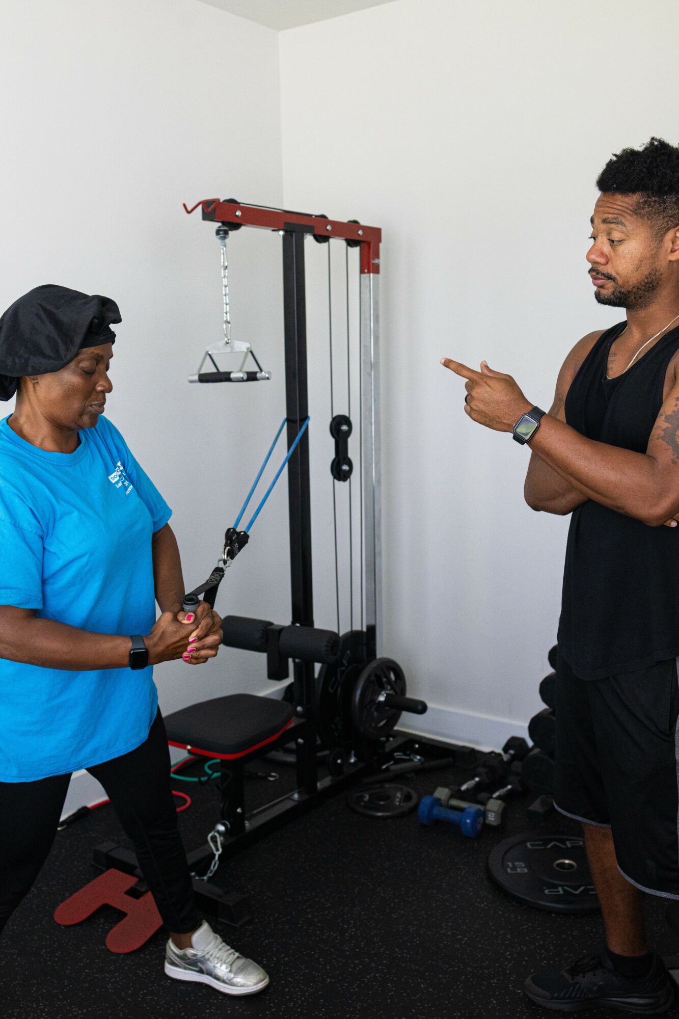A woman in a blue shirt and black cap uses resistance bands with a trainer in a gym, white wall background.