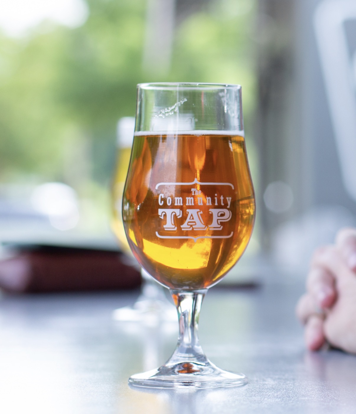 A glass of amber-colored beer with a logo reading 'Community TAP' on it, on a table with a blurred background.