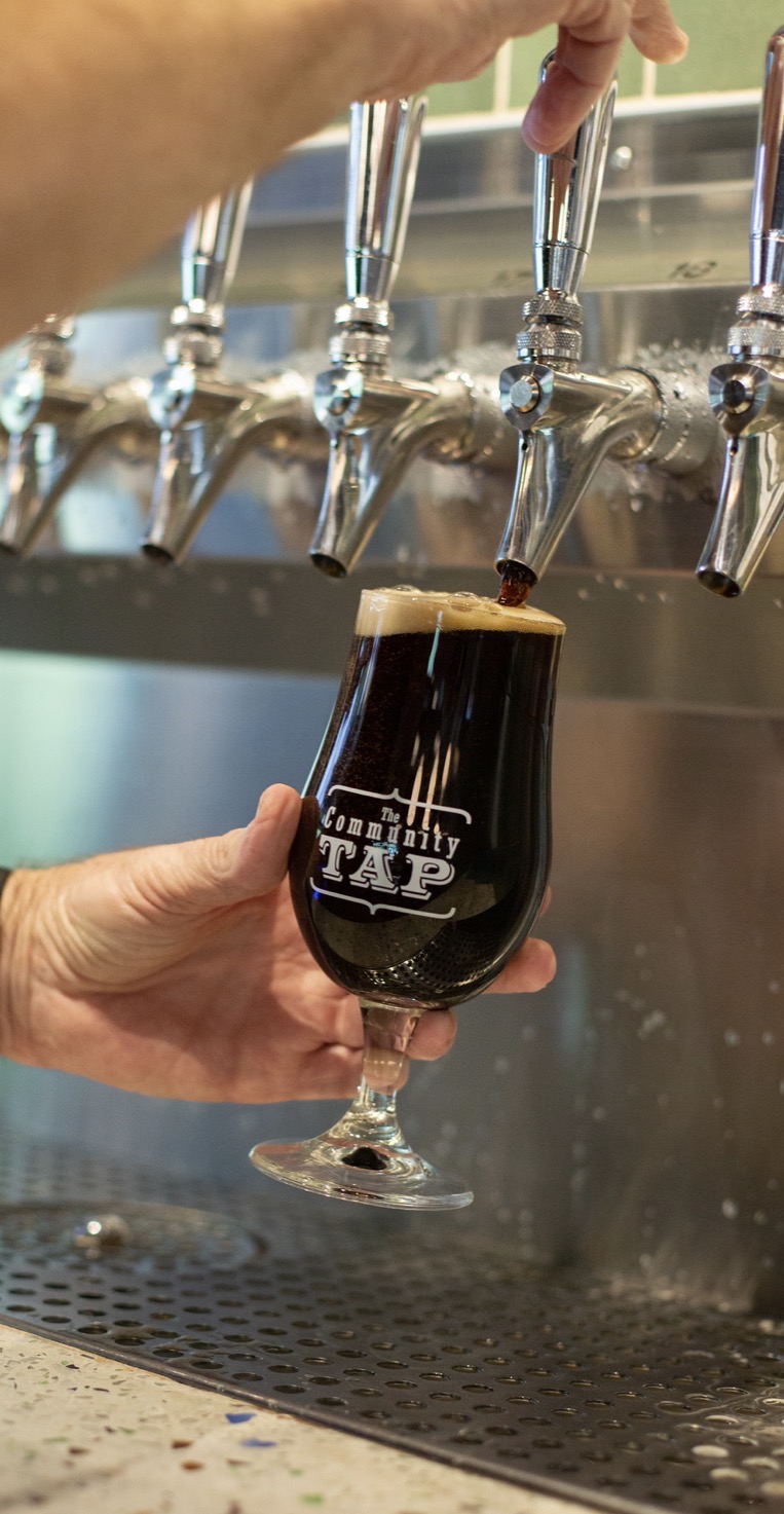 Person fills a glass with dark beer from a tap, with multiple taps visible in the background.