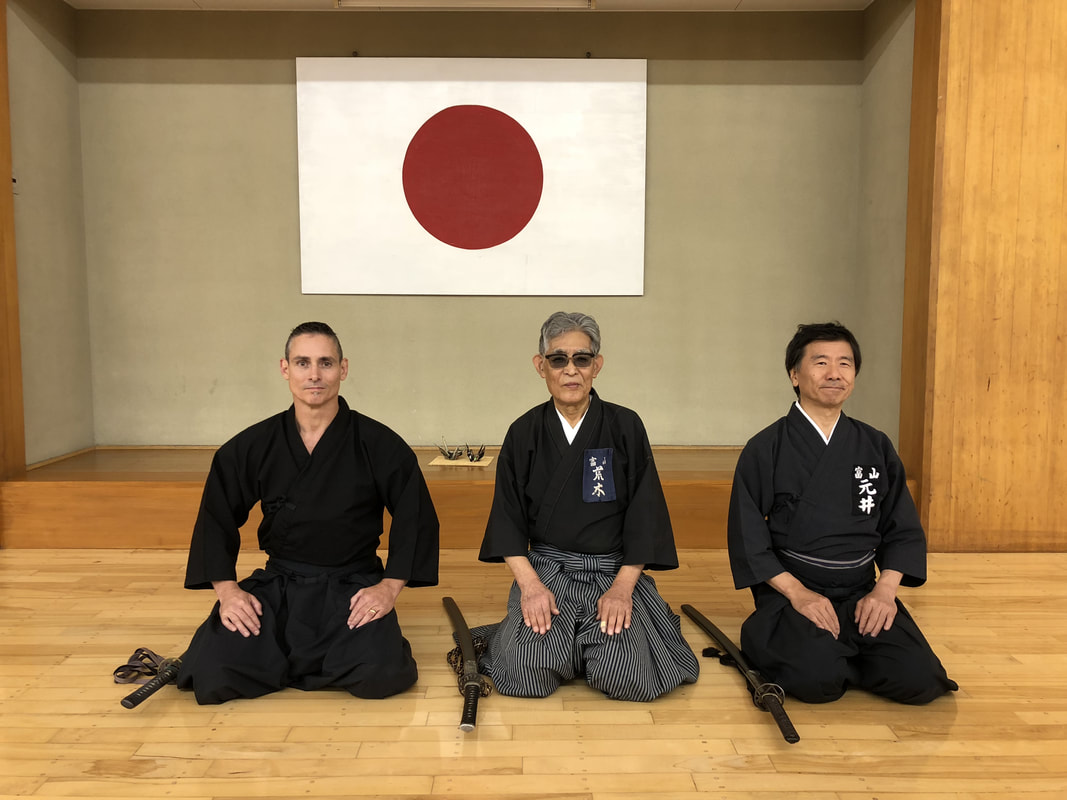Three people kneeling in traditional martial arts uniforms in front of a Japanese flag, indoors. All wear black uniforms with belts and weapons.