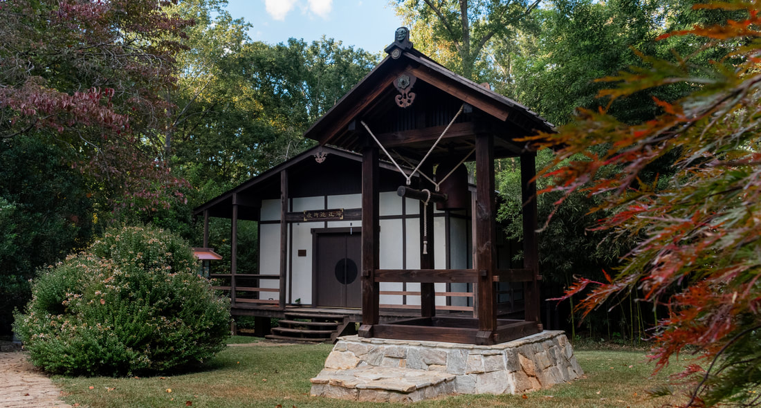 Small wooden building with a gabled roof, surrounded by trees and bushes, on a stone foundation.