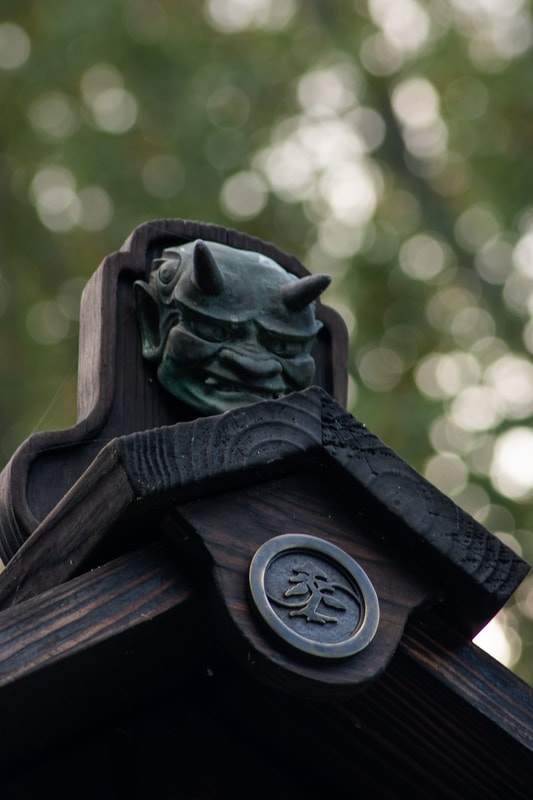 Close-up of a dark wooden structure with a small demon-like figure at the top, against a blurred green background.