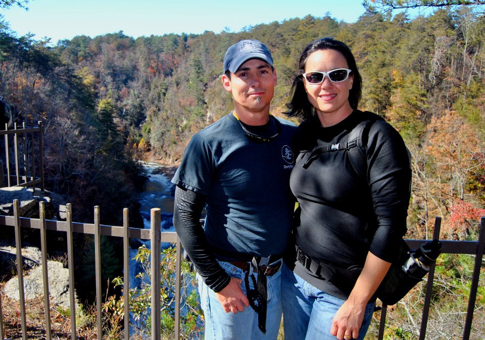 A man and woman stand outdoors near a railing with trees and a river in the background, smiling at the camera.