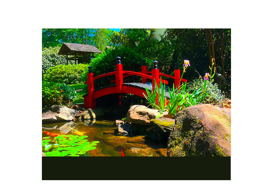A small red bridge over a pond with rocks, plants, and trees in a garden setting.