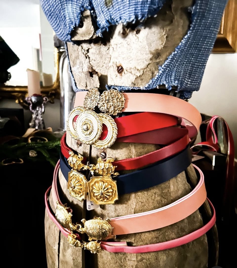 Display of multiple colorful belts with decorative buckles on a stone bust, with a blue fabric top and background objects.