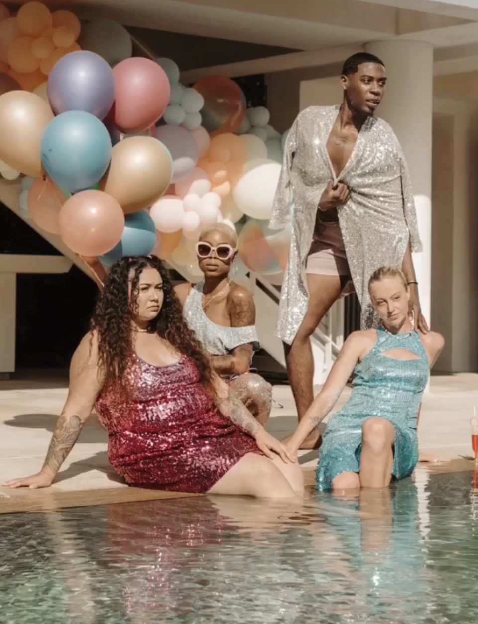Four people by a pool with colorful balloons in the background, one standing and three sitting or kneeling, wearing stylish clothing.