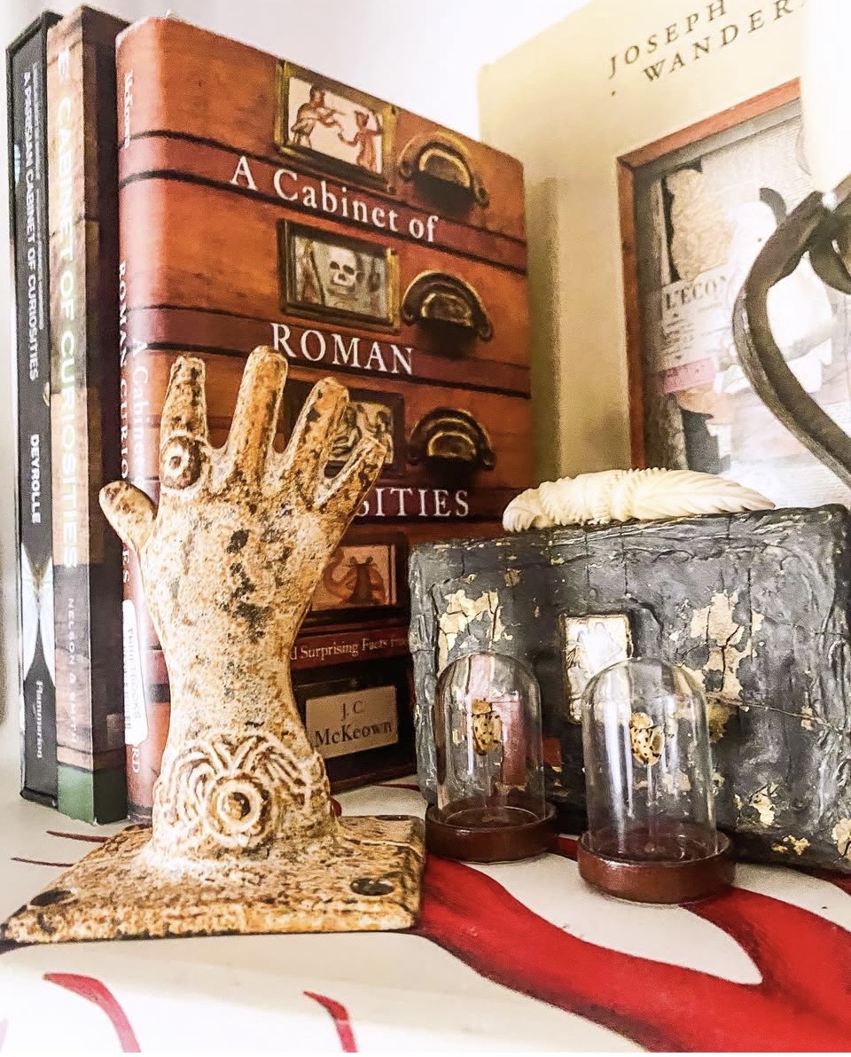 Decorative hand sculpture, books, and glass jars on a table with a red cloth, and a book titled 'A Cabinet of Roman' in the background.