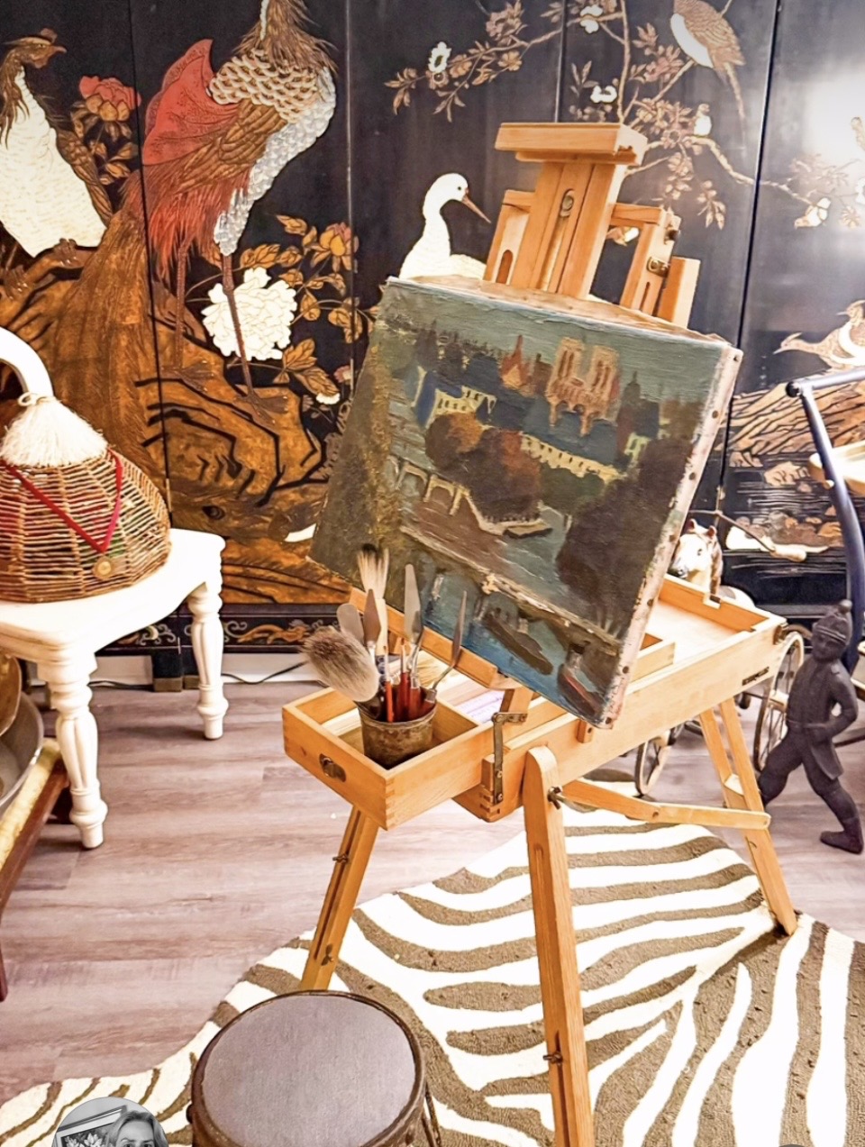 Easel with a landscape painting in a room with decorative wall art, chairs, and a striped rug.