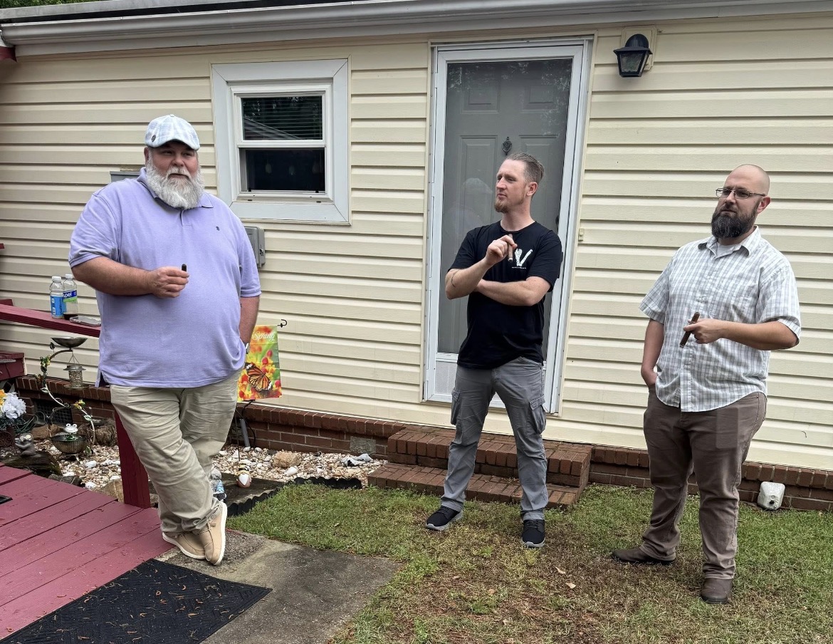 Three men standing outside a house, one with a beard and glasses, another with arms crossed, and the third holding a drink.
