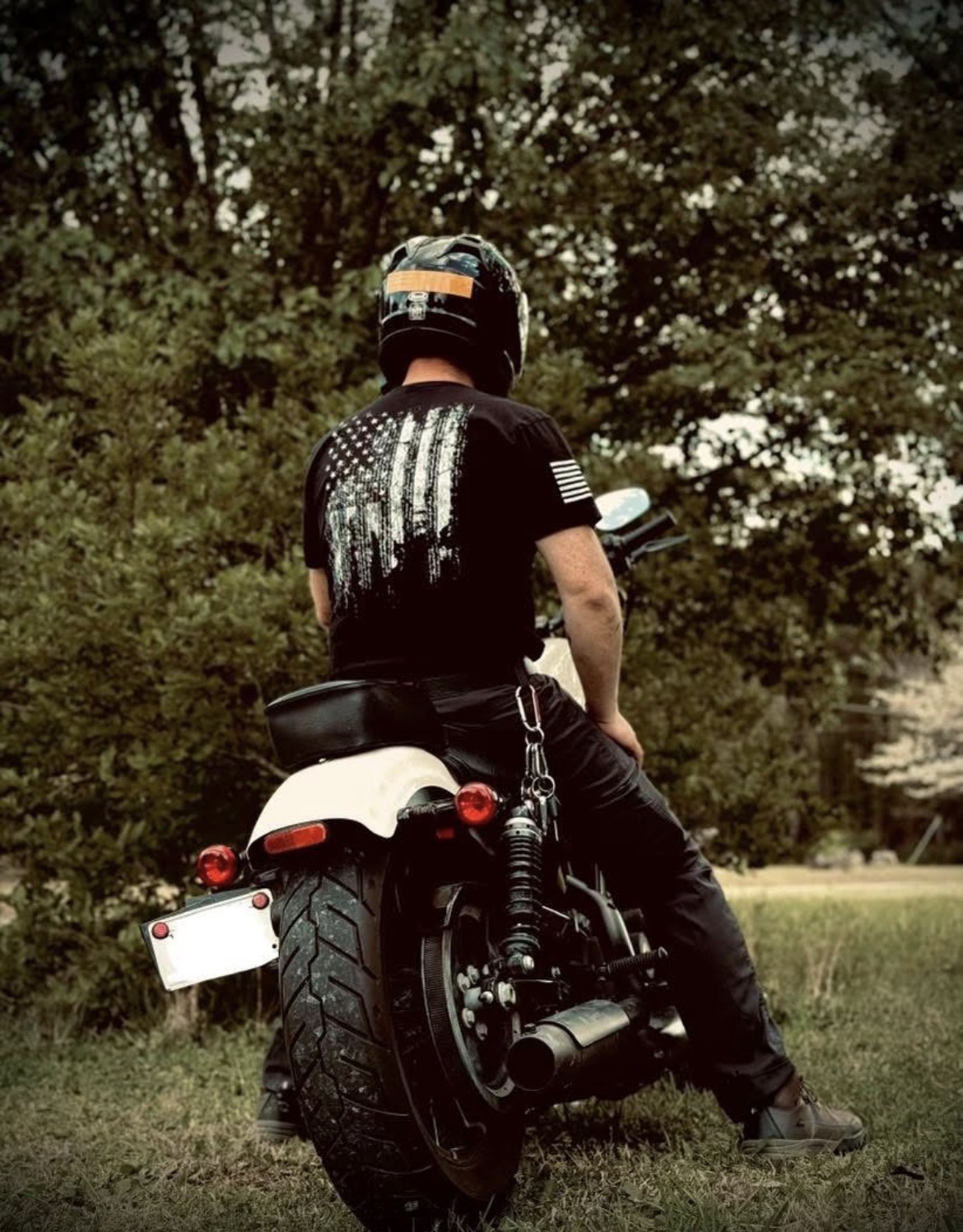 Person sitting on a motorcycle outdoors, wearing a helmet and black clothing, with trees in the background.