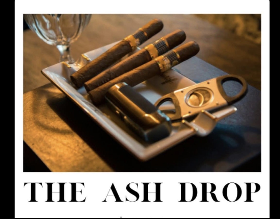 Cigars and a cigar cutter on a white plate with a glass of wine in the background, titled 'The Ash Drop'.