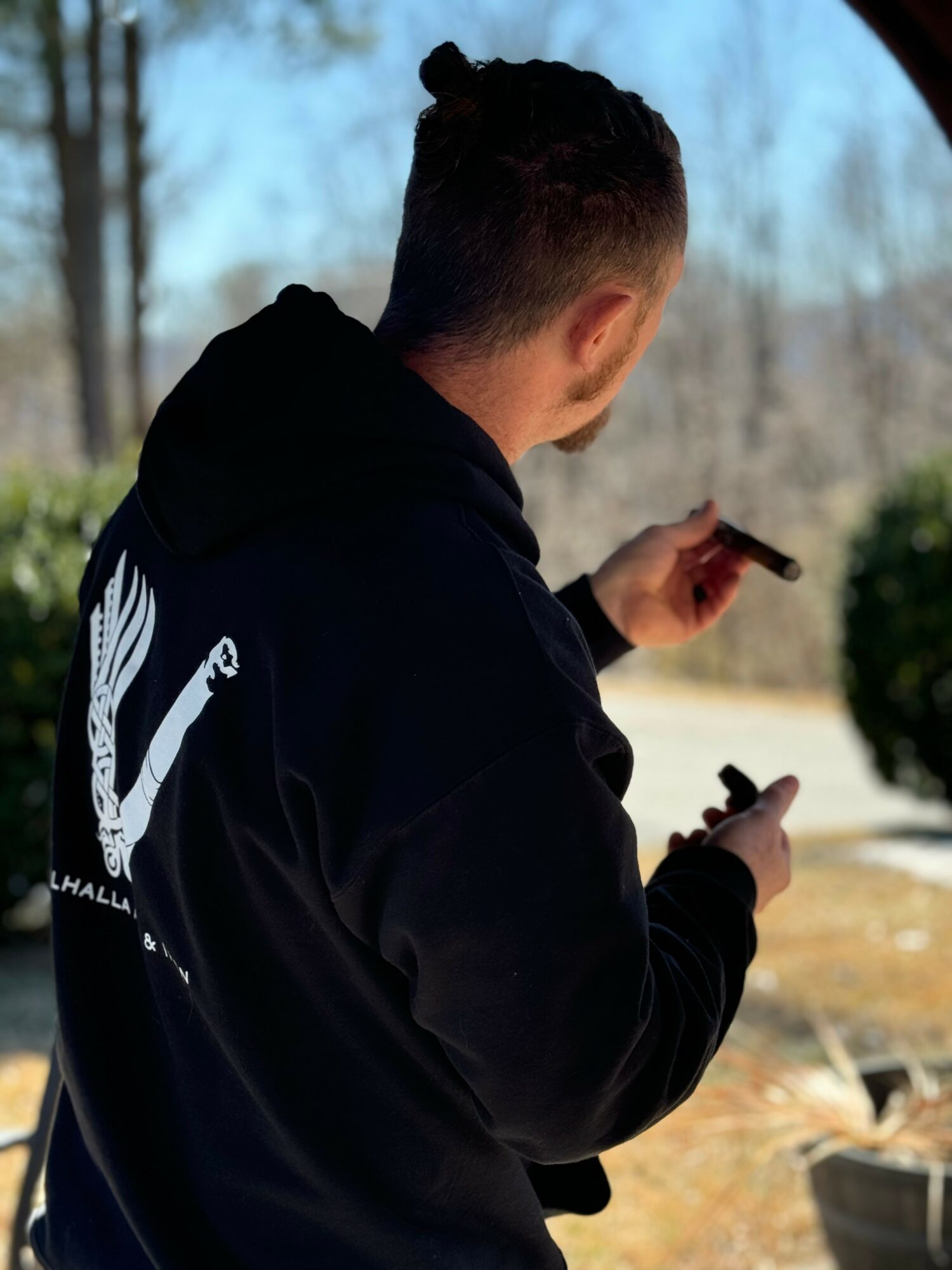 Person with short hair in a black hoodie looking at a phone outdoors, trees and bushes in background.