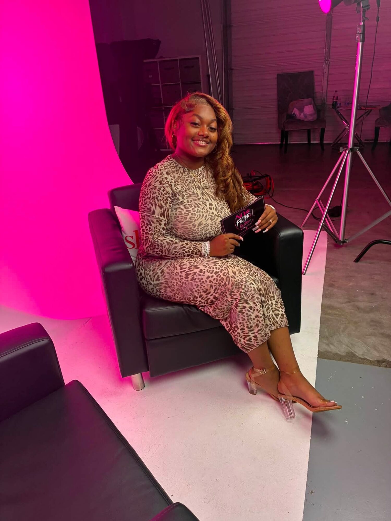 Woman sitting on a black chair, smiling, holding a clutch, wearing a leopard print dress, in a studio with pink lighting.